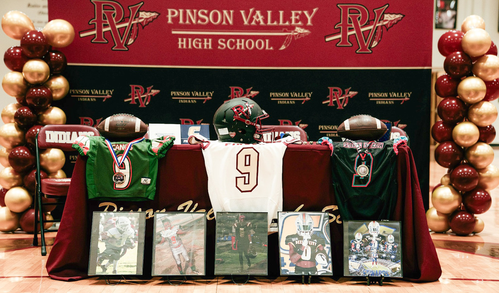 A table and background are highly decorated for national signing day with jerseys, balloons, photos, etc. The student athlete has his number nine displayed prominently along with pictures of his accomplishments. The scene is decorated with the Pinson Valley High School logo and colors of crimson and gold.