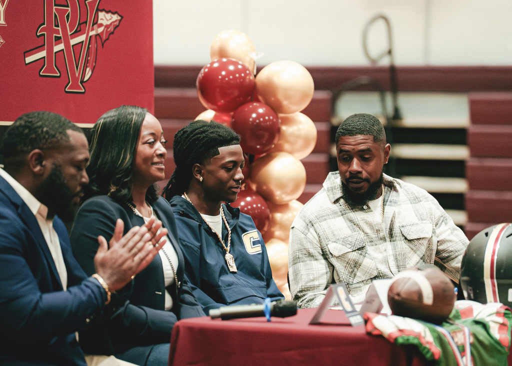 A family of three is sitting behind a decorated table at Pinson Valley High School on national signing day. The student's coach is sitting on the far left-hand side, clapping his hands.