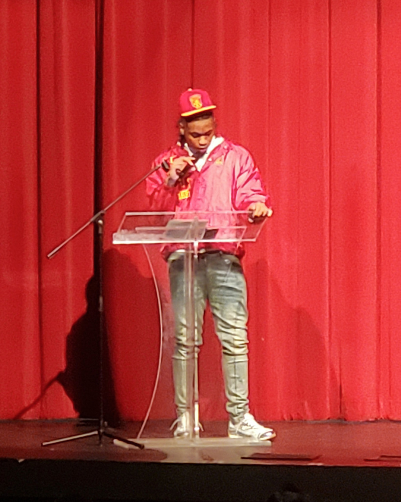 A Pinson Valley high school student stands behind a podium and speaks into a microphone during national signing day. There is a bright red curtain behind him.
