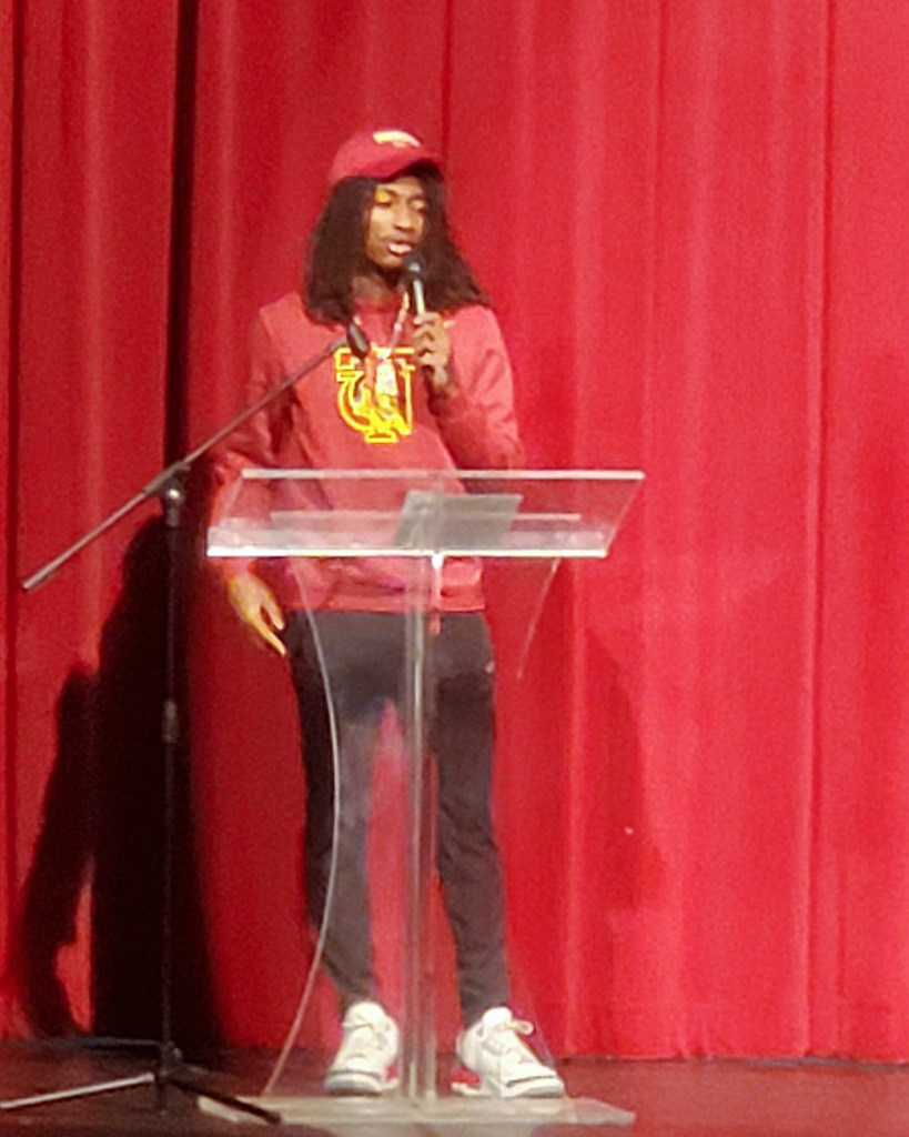 A Pinson Valley high school student stands behind a podium and speaks into a microphone during national signing day. There is a bright red curtain behind him.