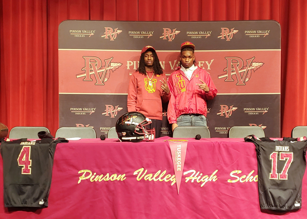 Two high school athletes stand behind a table during national signing day. They are holding up celebratory hand signals. The table and background are decorated with Pinson Valley High School logos and the colors of crimson and gold. The two students are wearing Tuskegee University jackets.