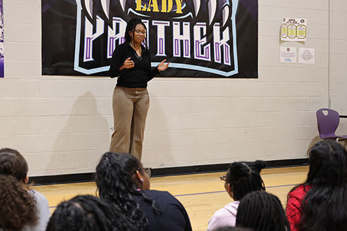 A woman speaks to a group of girls inside a school gym.