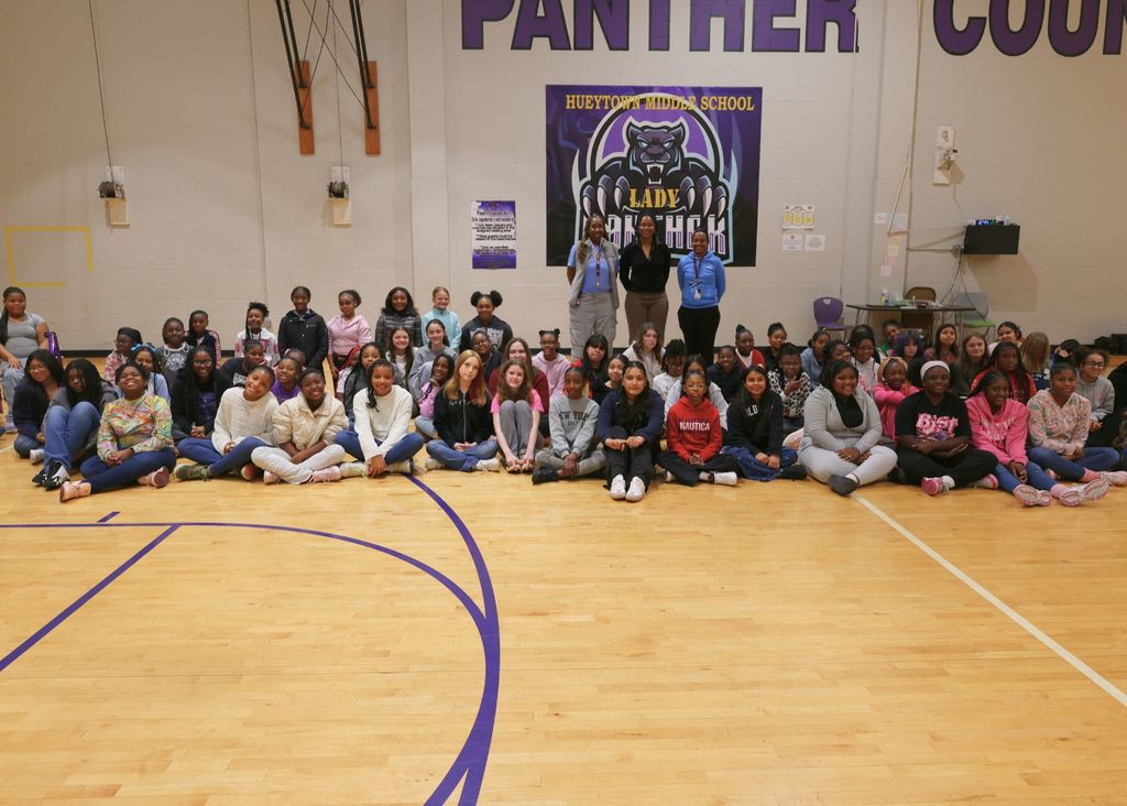 A large group of girls pose for a photo with three adult women inside a school gym.