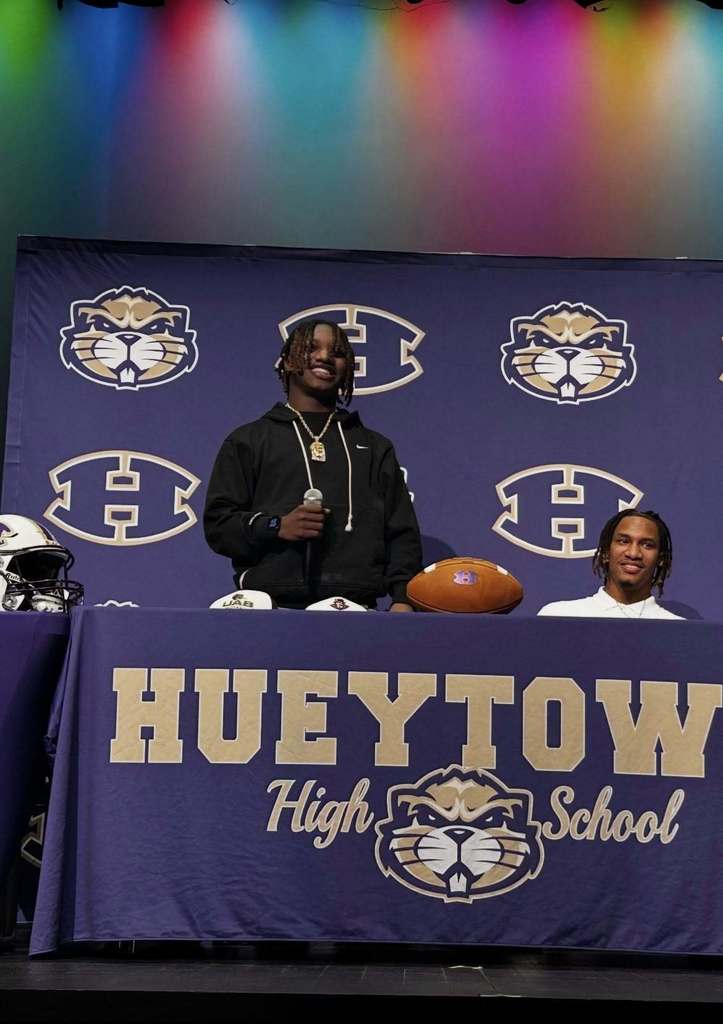 A high school football player stands behind a table and holds a microphone during National Signing Day. The table and background are decorated in Hueytown  High logos and colors of purple and gold. 