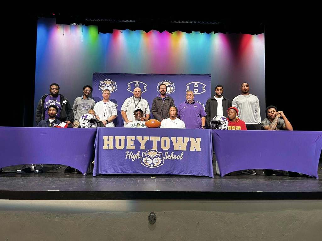 Photo from Hueytown High School on National Signing Day. A family is sitting behind a table decorated with the Hueytown high school logo and colors. There is also a purple and gold Hueytown banner in the background. Football helmets and a football are on the table for decoration.