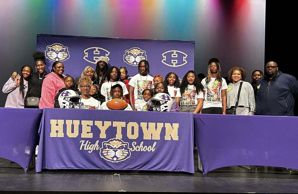Photo from Hueytown High School on National Signing Day. A family is sitting behind a table decorated with the Hueytown high school logo and colors. There is also a purple and gold Hueytown banner in the background. Football helmets and a football are on the table for decoration.