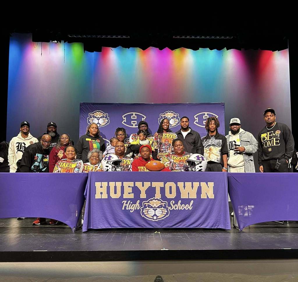 Photo from Hueytown High School on National Signing Day. A family is sitting behind a table decorated with the Hueytown high school logo and colors. There is also a purple and gold Hueytown banner in the background. Football helmets and a football are on the table for decoration.