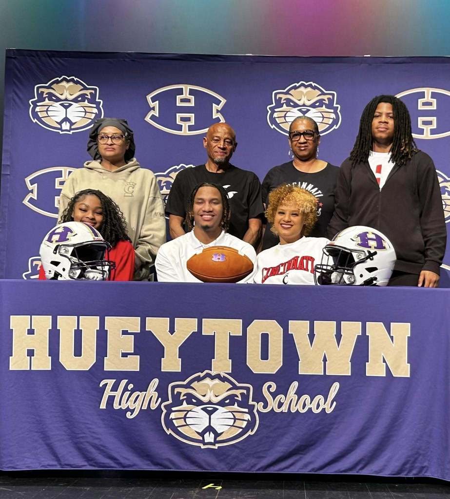 Photo from Hueytown high school on national signing day. A family is sitting behind a table decorated with the Hueytown high school logo and colors. There is also a purple and gold Hueytown banner in the background. Football helmets and a football are on the table for decoration.