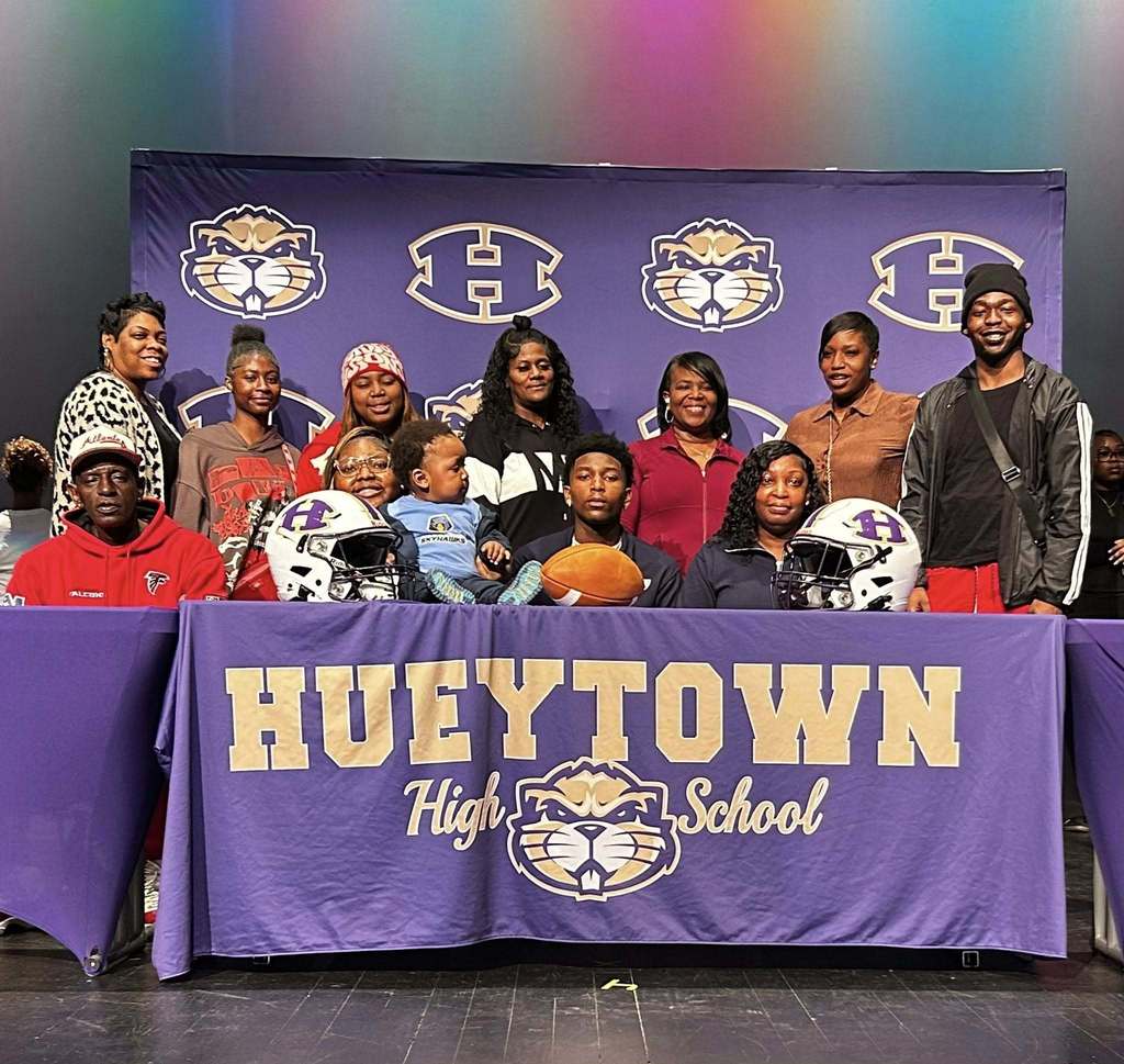 Photo from Hueytown High School on National Signing Day. A family is sitting behind a table decorated with the Hueytown high school logo and colors. There is also a purple and gold Hueytown banner in the background. Football helmets and a football are on the table for decoration.