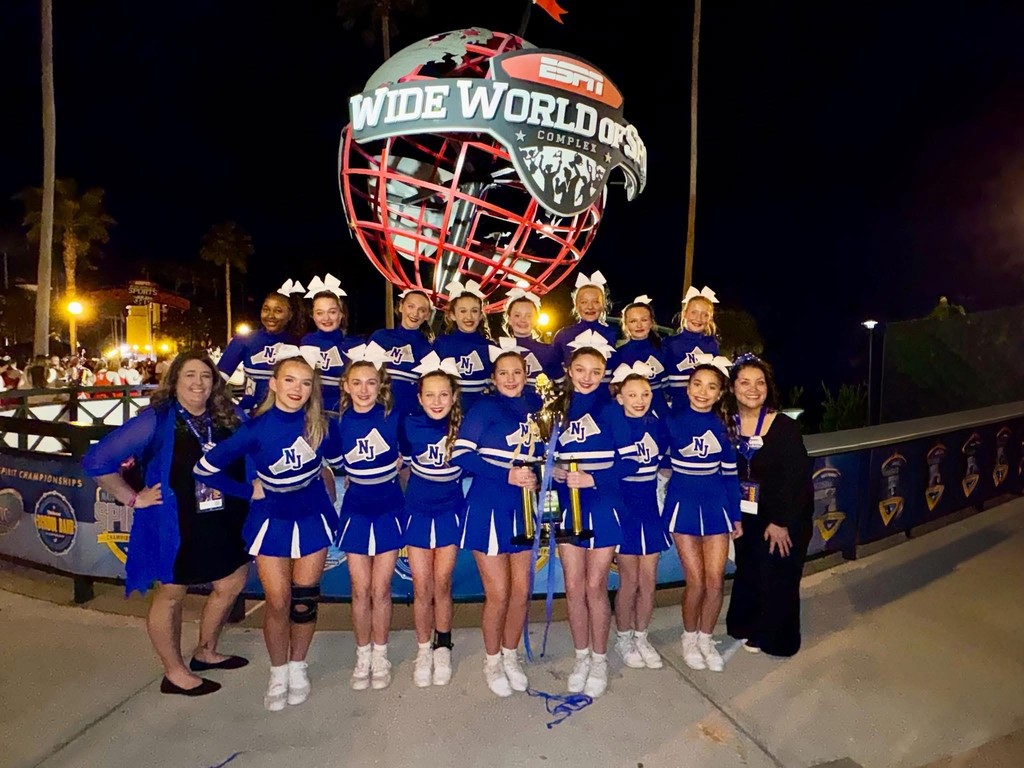 North Jefferson Middle School cheerleaders stand for a photo with their coaches outside in front of a structure shaped like a globe that says ESPN Wide World of Sports. The cheerleaders hold a trophy.