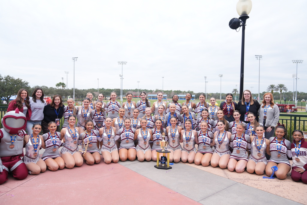 Gardendale High School cheerleaders and dance team members pose for a photo with their coaches and the mascot. They all have their uniforms on and are wearing medals. In front of the members is a trophy.