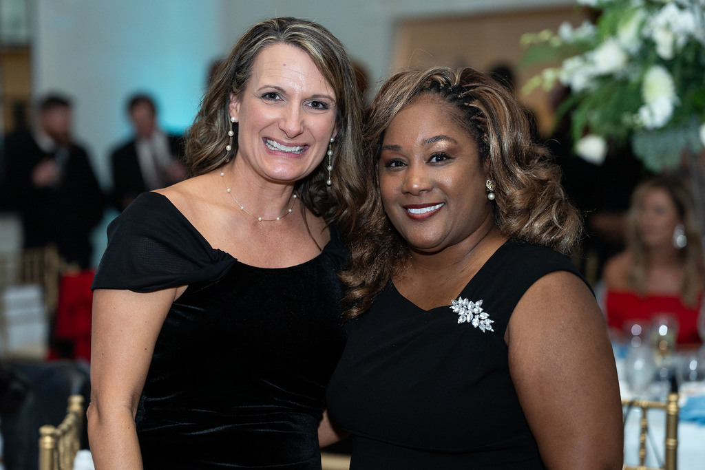 Two women stand together for a photo and smile. Both of the women are wearing black formal dresses.