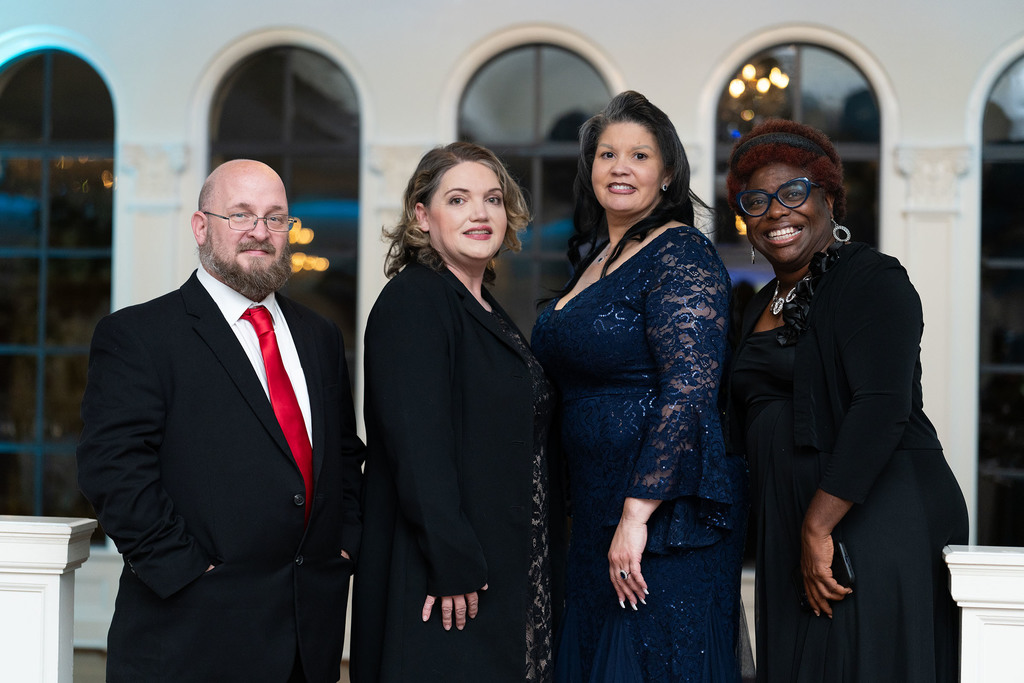 A man and three women stand together and smile for a photo. The man is in a suit and the women wear formal dresses.
