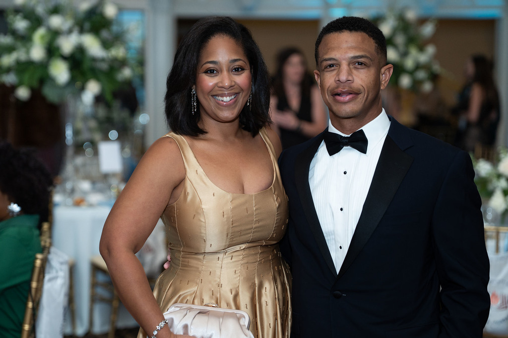 A man and a woman stand together for a photo and smile. The woman is in a gold formal dress and the man is in a tuxedo.