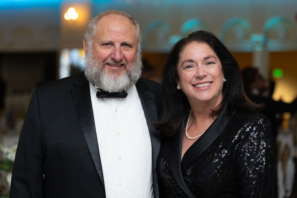 A man and a woman stand together for a photo and smile. The main is in a tuxedo and the women wears a sequin suit jacket.