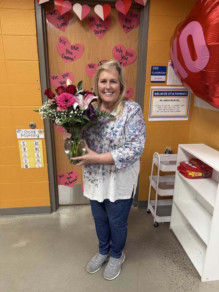 A school counselor is being celebrated during national school counselor appreciation week. She is holding a vase full of pink and red flowers. In the background are several cut out red hearts with custom messages.