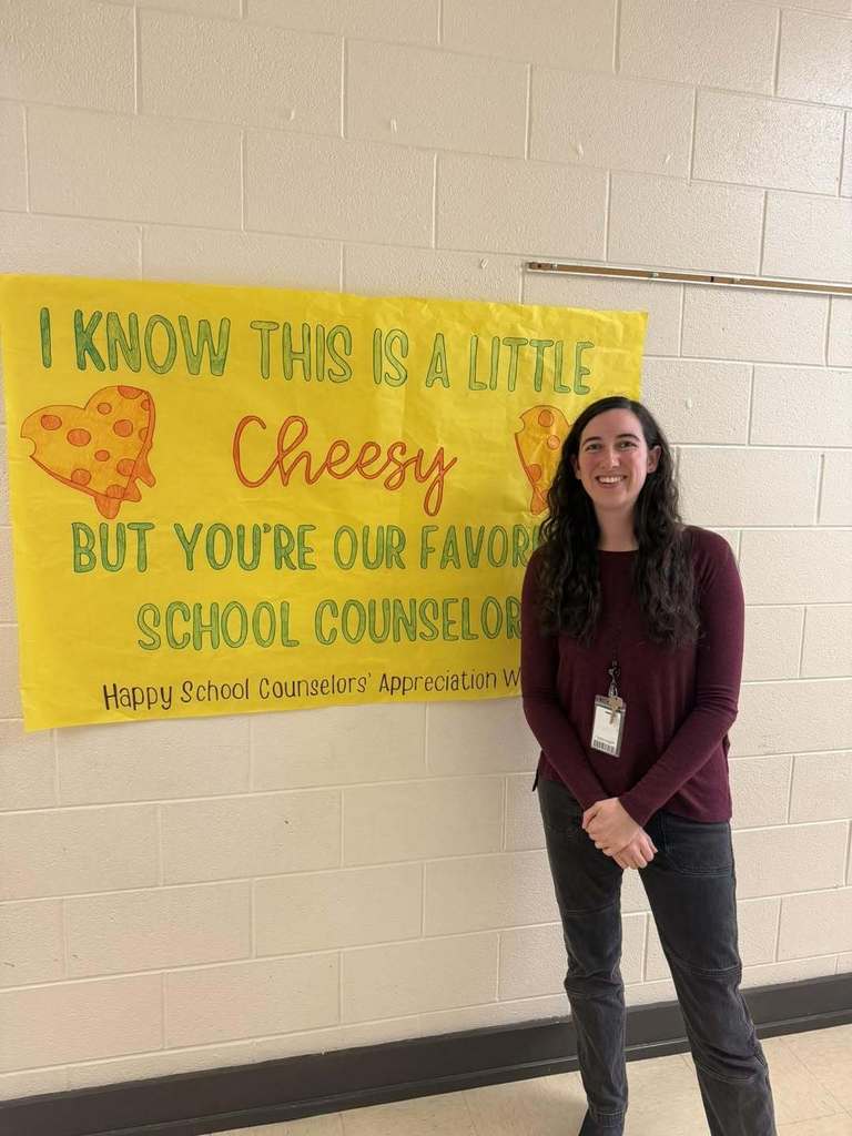 School counselor is standing in front of a banner that says, "I know this is a little cheesy, but you're our favorite school counselor. Happy school, counselor appreciation week."