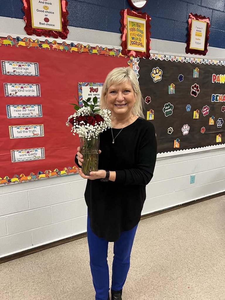 A school counselor is standing in an elementary school classroom. She is holding a vase full of red roses. She is being celebrated for national school counseling week.