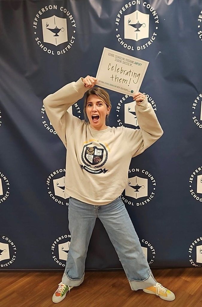 A school counselor stands in front of the Jefferson County school district background. She is celebrating and holding her arms up. The sign in her hands says school counseling programs amplify student success by celebrating them!