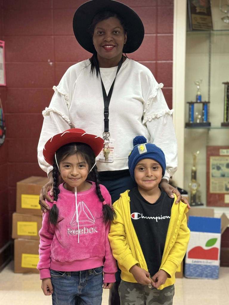 A school counselor stands with two young elementary school students. They are all wearing unique hats. The school counselor in the back has on a black cowboy style hat, the girl in the front left is on a red, cowboy hat, and the boy on the right has on a blue beanie style hat. 