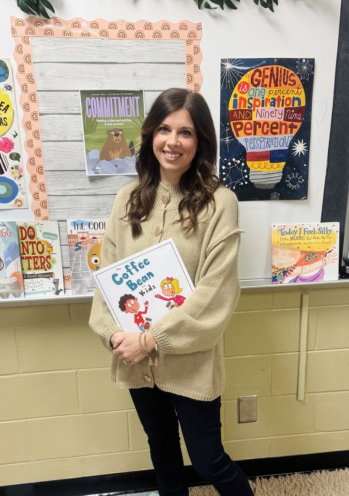 A school counselor is standing in front of a colorful bulletin board. She is smiling and holding a book that says the coffee bean for kids.