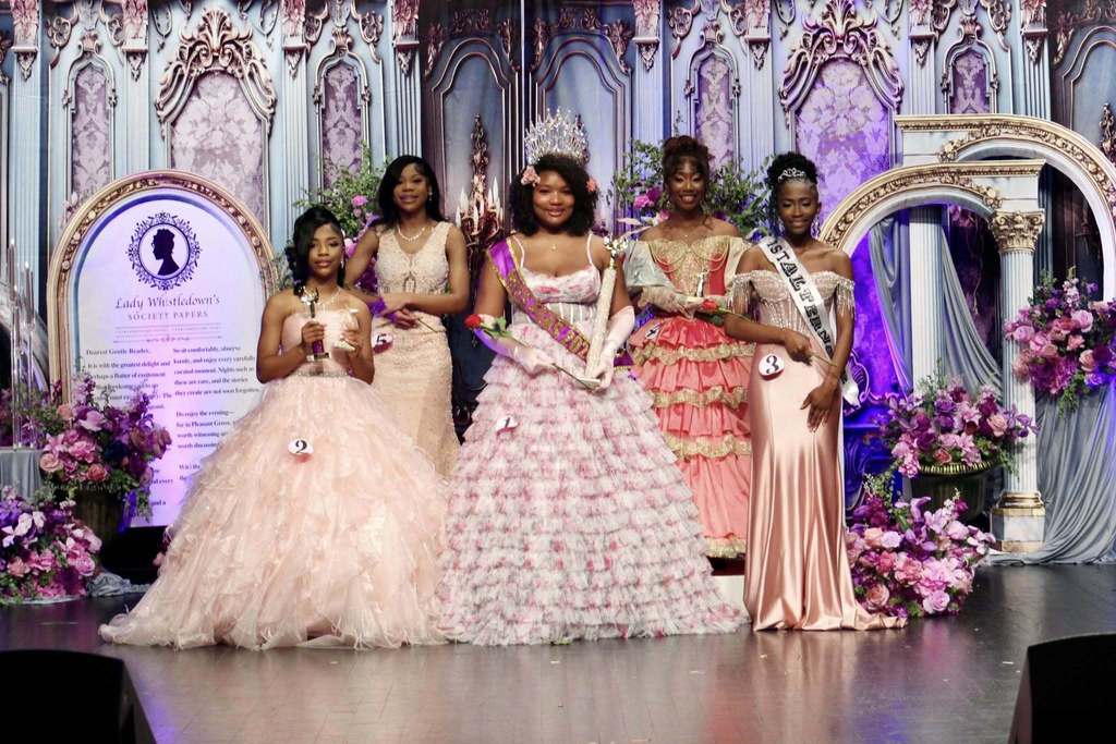 Young ladies wear ballgowns and smile while standing on a stage decorated in Bridgerton theme. The girls smile and hold awards/wear sashes. The girl in the middle wears a sash, holds a trophy and wears a crown.