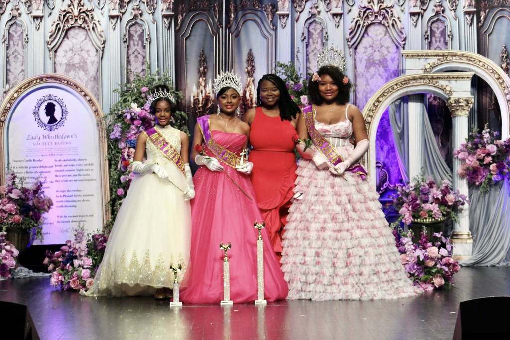 Three girls and a woman stand together for a photo. The girls hold trophies and roses and wear sashes and crowns. The woman standing with them is wearing a red dress. The four of them stand on a stage decorated in Bridgerton theme.