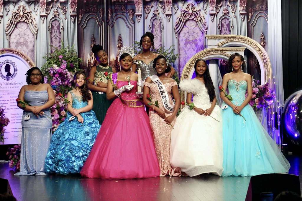 Girls stand together for a photo and wear ballgowns on a stage decorated in Bridgerton theme. Some of the girls hold roses, and two girls wear sashes. One of these girls also has a crown on her head and a trophy in her hand.