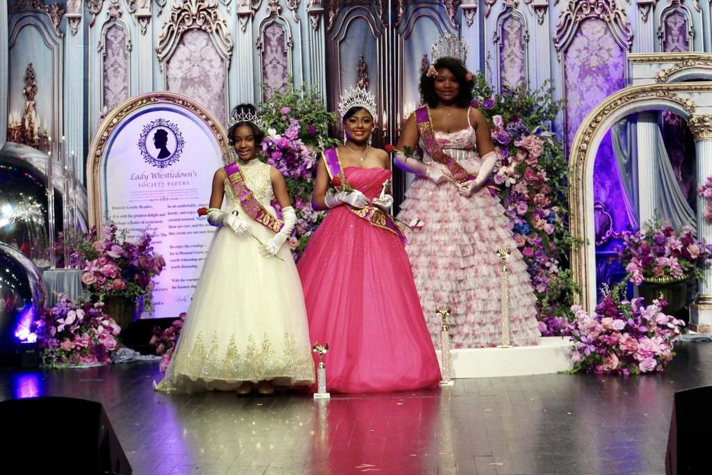 The winners of the Miss Pleasant Grove Elementary School, Middle School and High School pageants wear ballgowns, sashes and crowns and stand together for a photo. They stand on a stage decorated in Bridgerton theme. 
