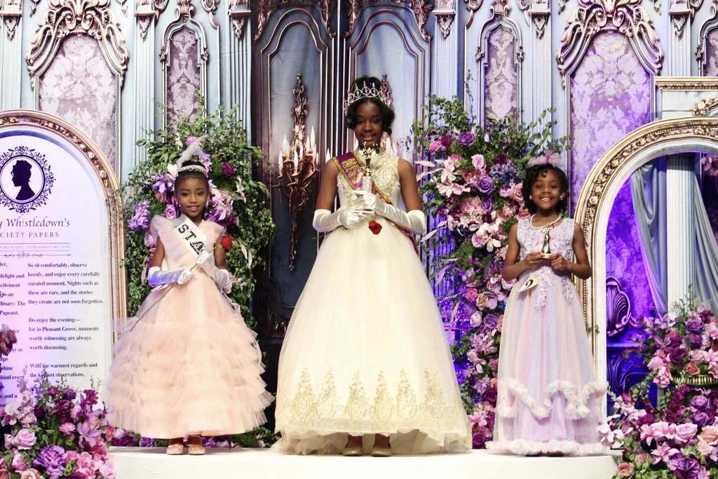 Three little girls stand together for a photo. They wear ballgowns and stand on a stage decorated in Bridgerton theme. One girl holds a rose and wears a sash, another girl holds a trophy, and the third girl holds a trophy, wears a sash, and has a crown on her head.