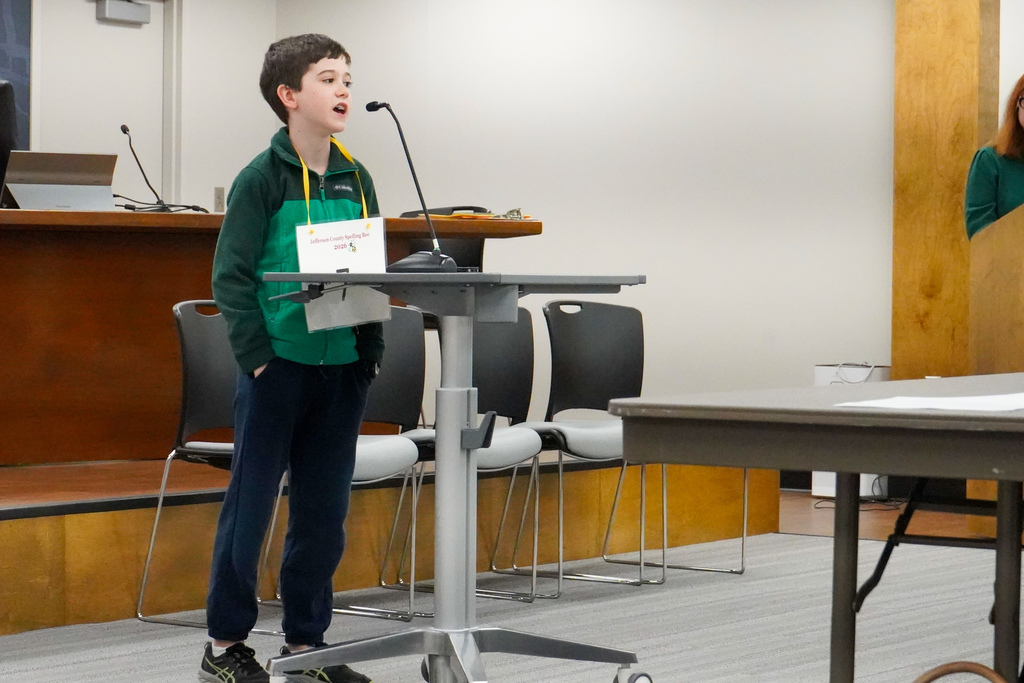 A student participating in the Jefferson County Spelling Bee spells a word into a microphone. The spelling bee is taking place inside the Jefferson County Schools board room.