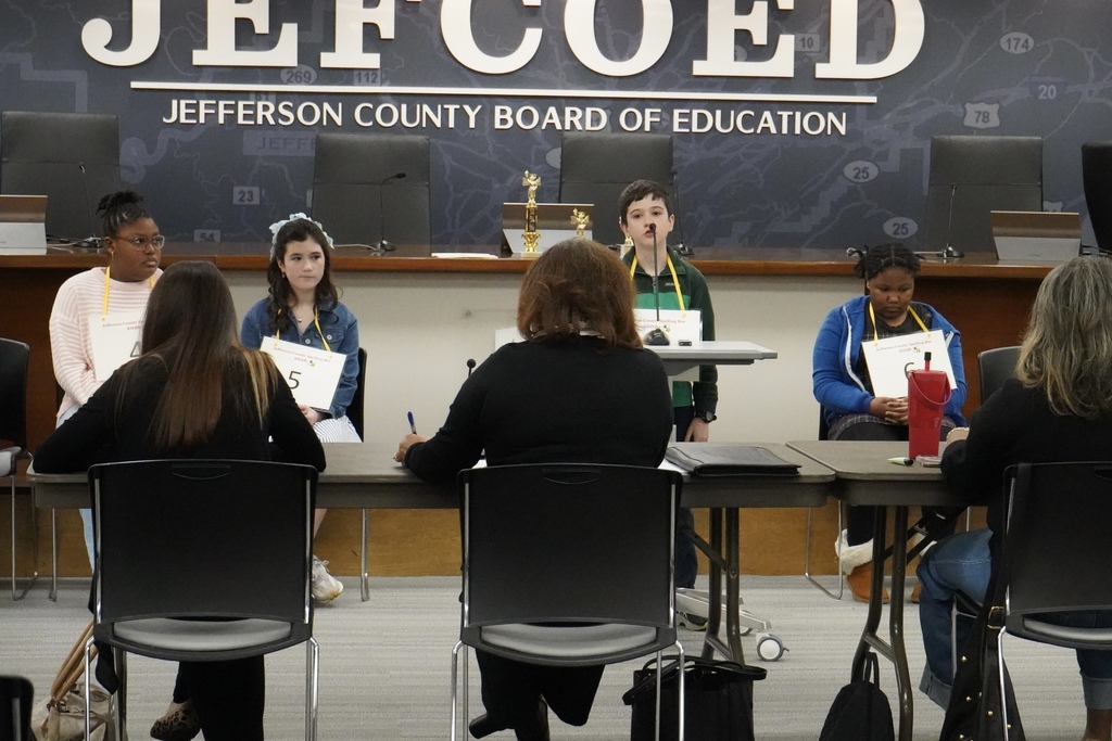 A student participating in the Jefferson County Spelling Bee spells a word into a microphone. Judges sit at a table in front of him and his fellow spellers sit in chairs behind him.