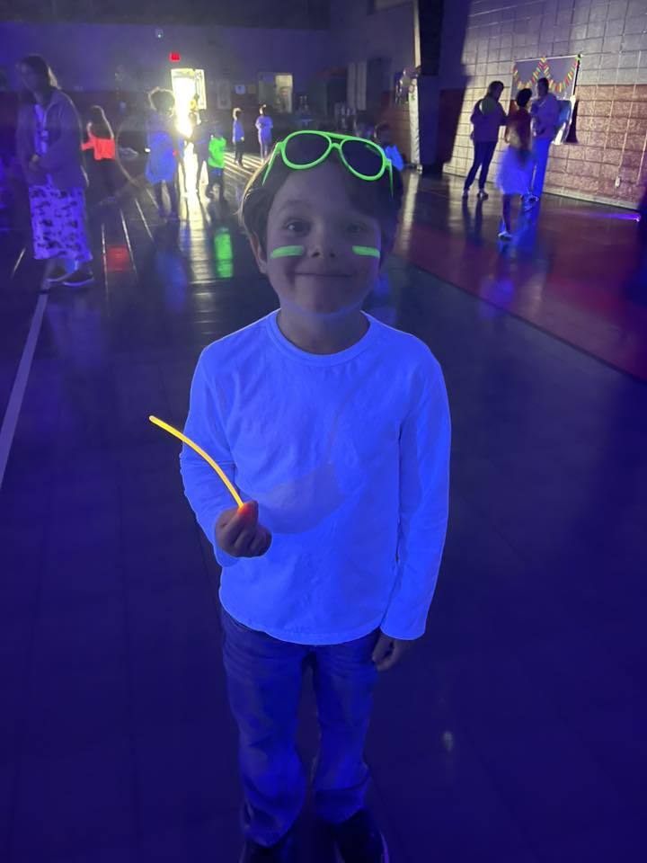 A little boy stands in a dark school gym and smiles. He wears glow-in-the-dark sunglasses on top of his head and holds a glow stick. He also has glow-in-the-dark face paint on his face.