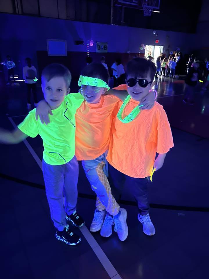 Three boys wearing glow-in-the-dark outfits stand together for a photo in a dark school gym and smile.