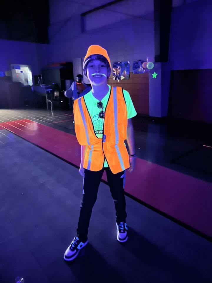 A boy wears a glow-in-the-dark shirt, vest, hat, and painted on mustache inside a dark gym during a glow party.