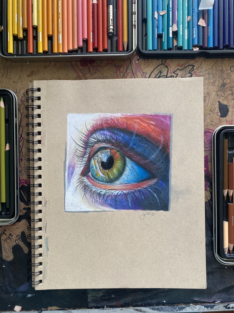 Photograph shows a drawing of a colorful eye on a notebook. The notebook is sitting on a table with colored pencils around it.