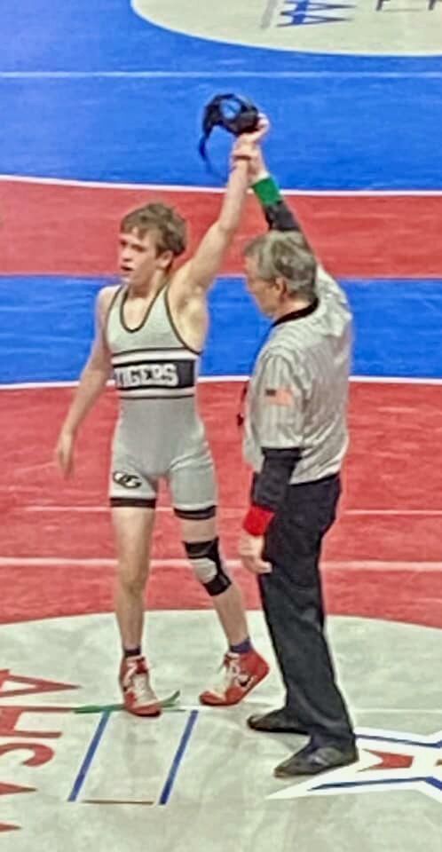 A wrestling official holds up Oak Grove High School wrestler David Hill's arm to show he was victorius during a wrestling match. David wears an Oak Grove wrestling uniform and the official wears a black and white striped shirt and black pants.