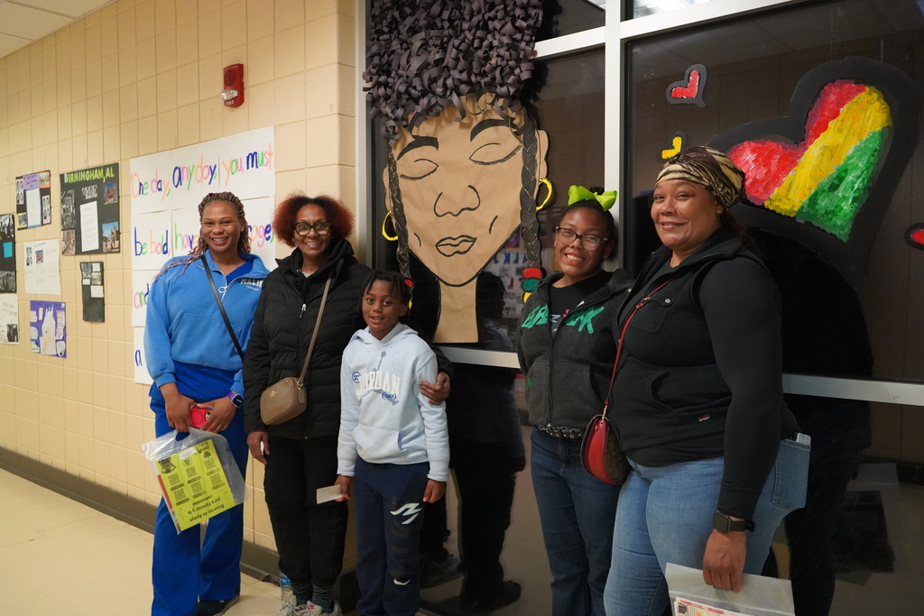 Three adults and two children stand together for a photo in a school hallway and smile. They stand in front of displays set up for Black History Month.