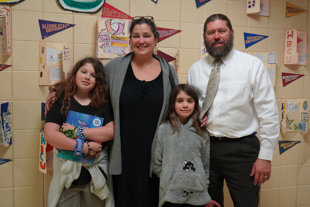 Two adults and two children stand together for a photo in a school hallway and smile. They stand in front of student-made presentations  highlighting HBCUs.