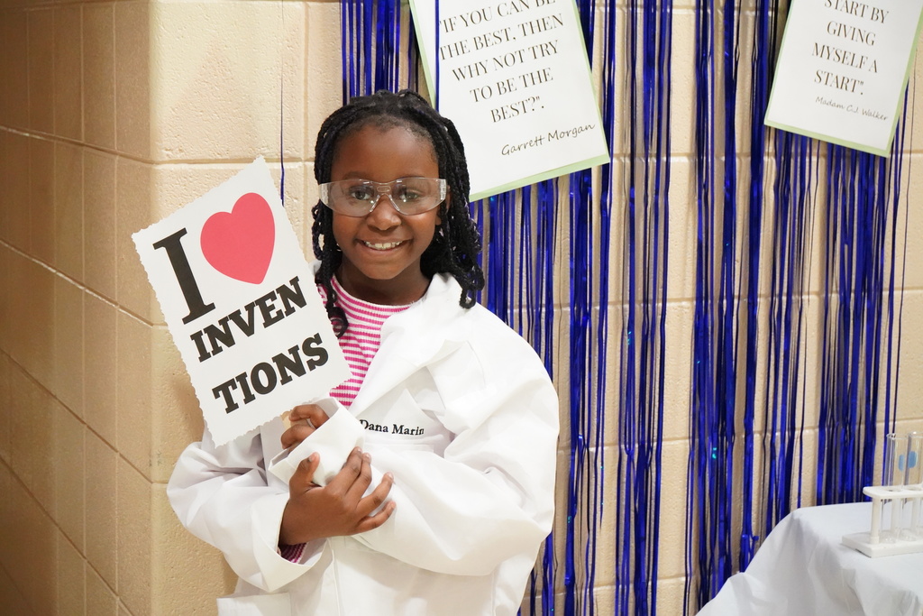 A little girl wears a lab coat and poses for a photo in front of a blue streamer backdrop. The little girl holds a sign that says "I heart inventions".