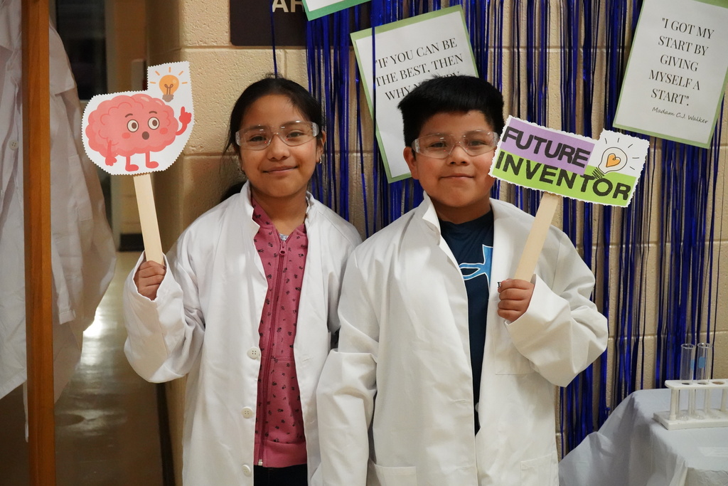 A girl and a boy stand together for a photo in front of a blue streamer photo backdrop. The students wear lab coats and hold up signs. One of the signs says "Future Inventor" and the other is an image of a brain with a lightbulb hovering over it.