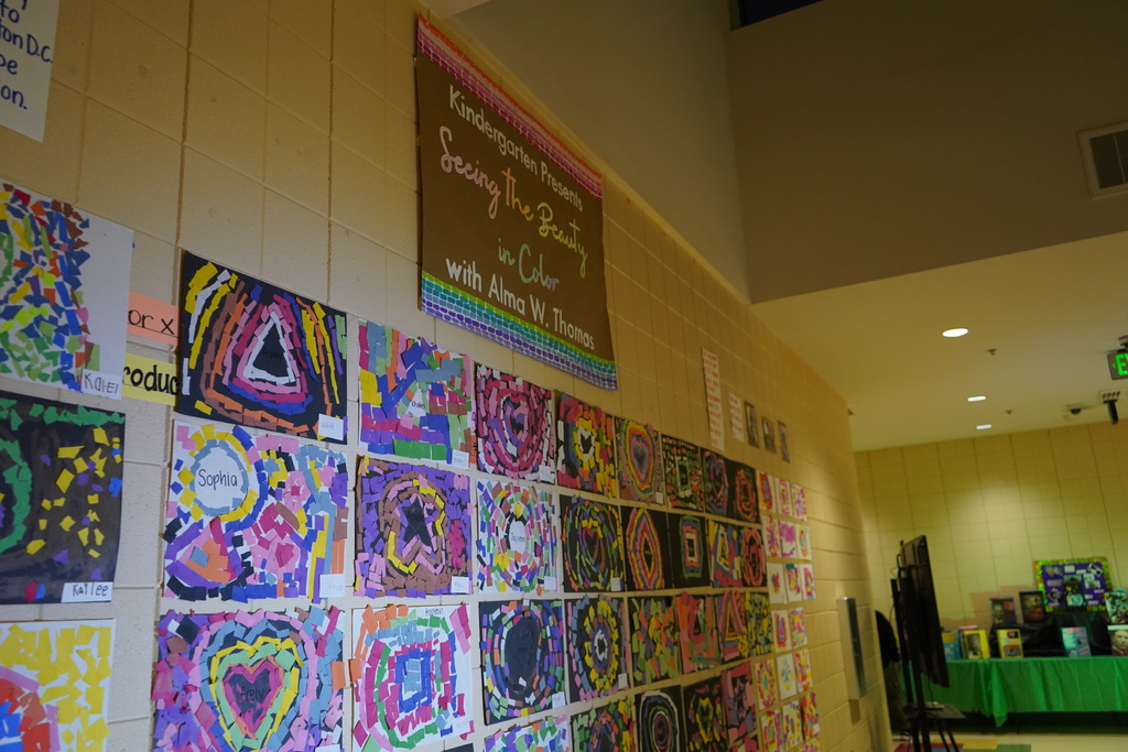 Student-created art made of colorful bits of paper are on display in a school hallway. Above the art is a sign that says "Kindergarten Presents Seeing the Beauty in Color with Alma W. Thomas."