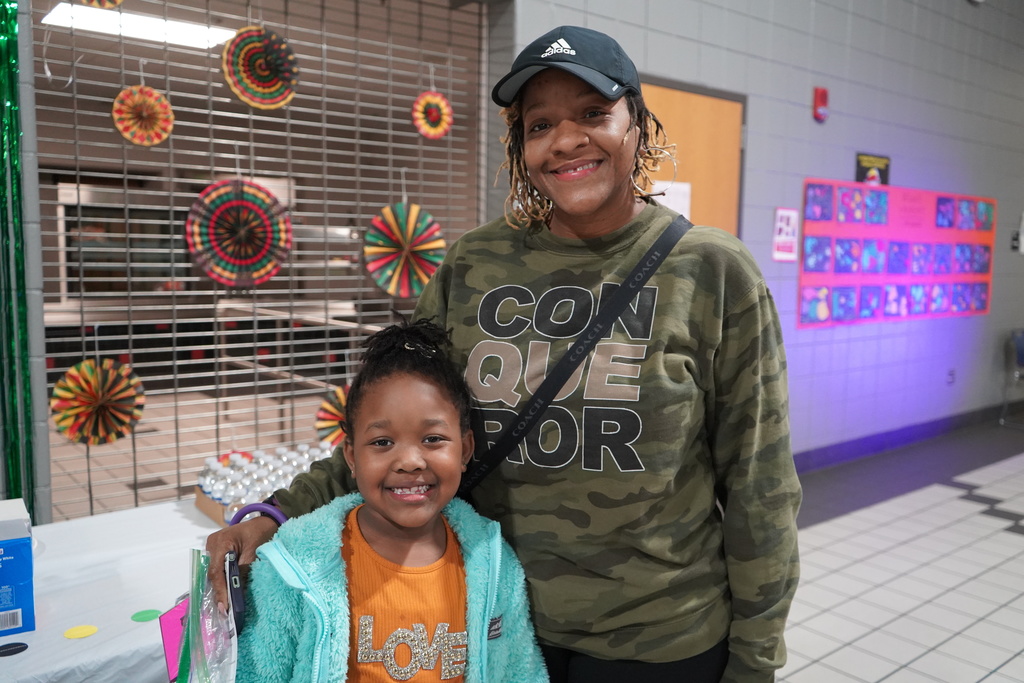 A woman and a girl stand together for a photo inside a school lunchroom. Both of them smile.