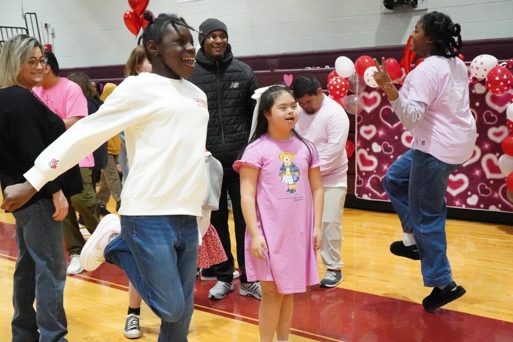 A group of students dance while attending a Valentine's Day themed dance. Adults stand in the background and smile.