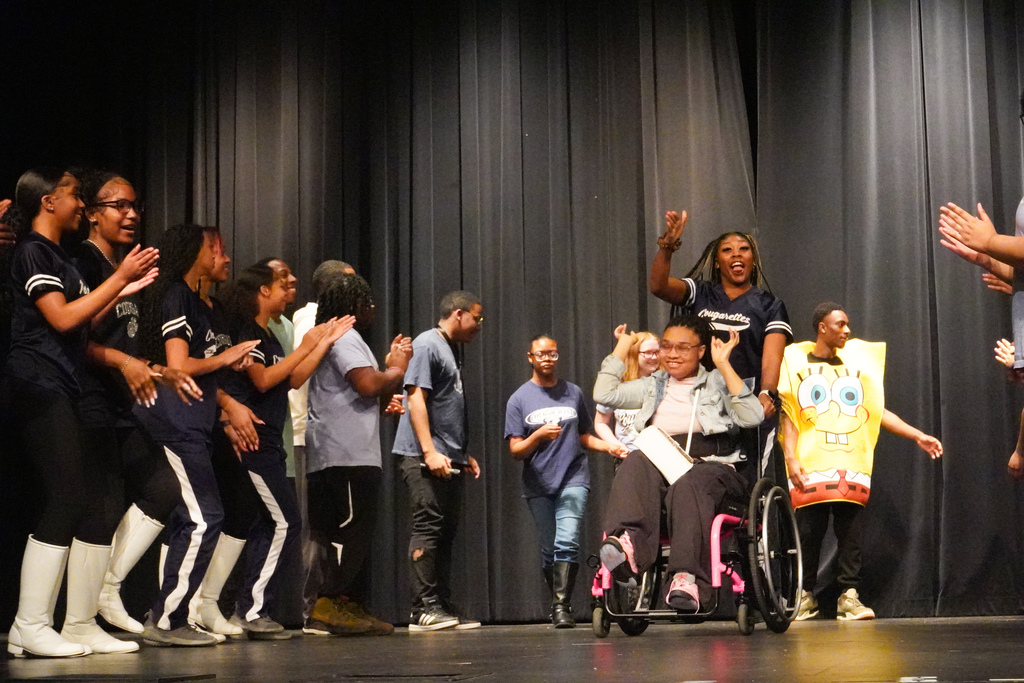 A group of students dance on a stage. Some of the students wear matching blue jerseys. One of the girls in a jersey pushes a girl in a wheelchair down the stage. A student dressed like Spongebob dances behind them. 