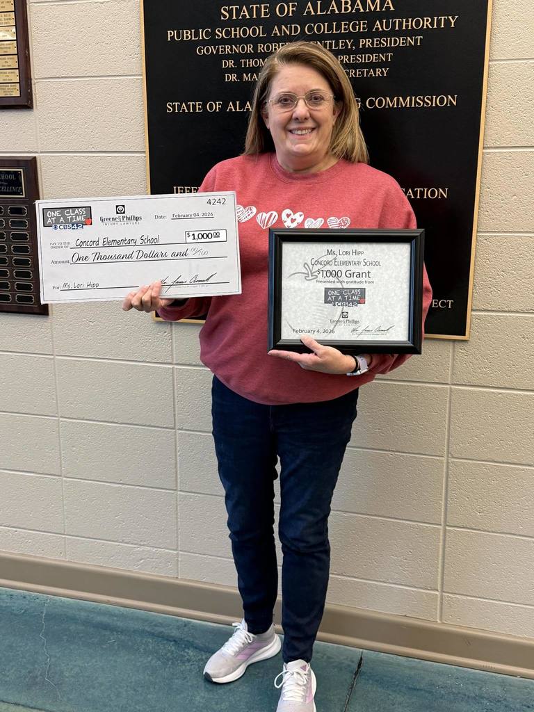 Concord Elementary School teacher Ms. Hipp stands in the school hallway and holds a framed piece of paper and a large check. The paper announces her as the recipient of a CBS 42 'One Class at a Time Grant'. The check is for $1,000.