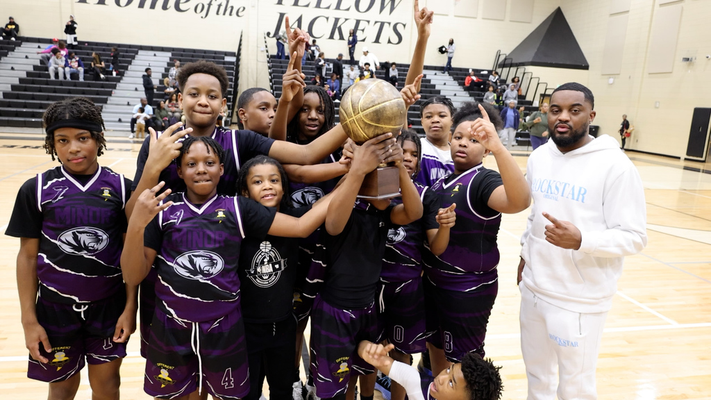 Minor Middle School's boys basketball team wear uniforms and pose for a photo with their coach in a school gym. They hold a trophy with a golden basketball on top. 