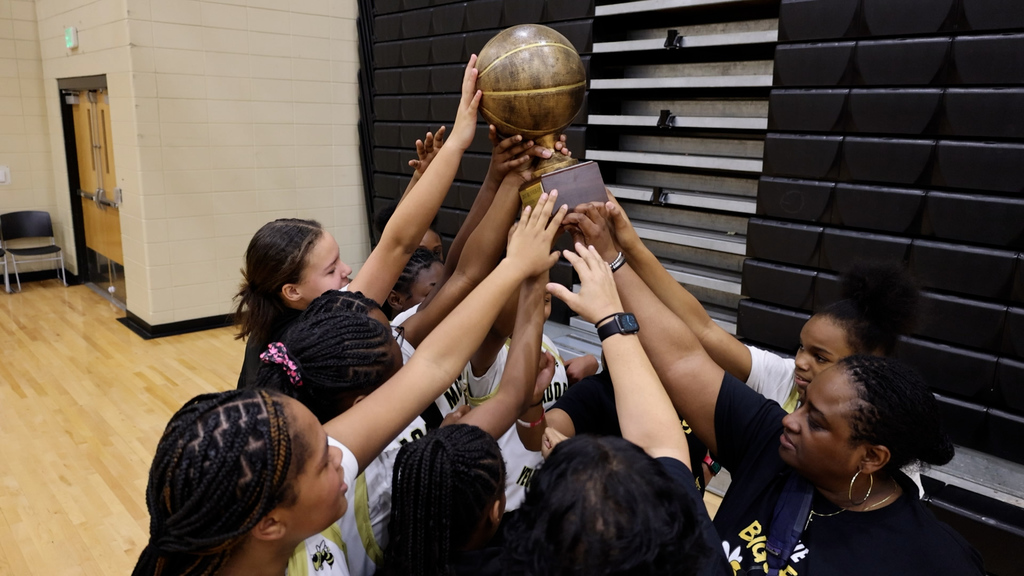 McAdory Middle School's girls basketball team holds a trophy in the air while standing in a school gym. The trophy is in the shape of a basketball.