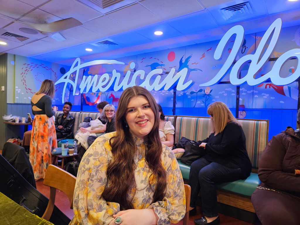 Alex Kanaday sits in a room with a big American Idol sign on the wall behind her and smiles for a photo. There are people in the room behind her sitting in booth style seats and standing up.