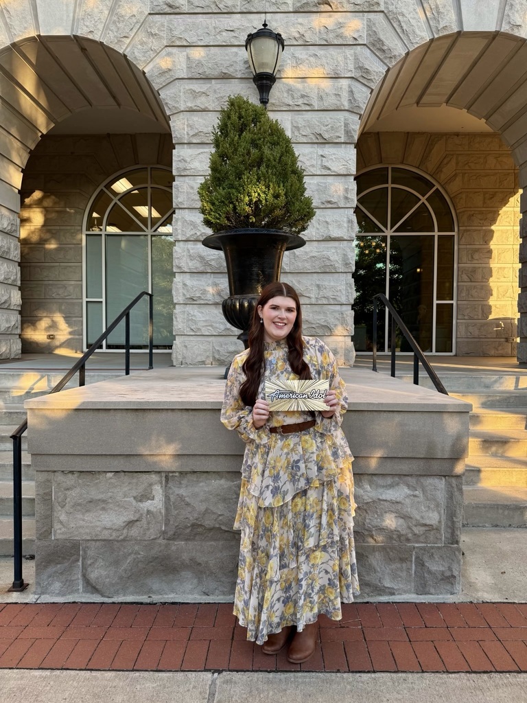 Alex Kanaday holds up an American Idol Golden Ticket and smiles for a photo. She stands outside a stone building.